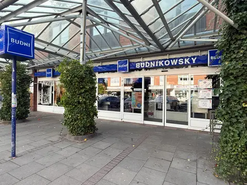 Eingang einer Budnikowsky-Drogerie unter einem Glasdach, davor ein gepflasterter Vorplatz mit efeubewachsenen Säulen und blauer BUDNI-Stele