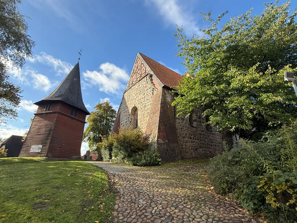 Roter Holzglockenturm mit Schieferspitze neben dem Fachwerkgiebel einer Feldsteinkirche, davor ein Kopfsteinpflasterweg