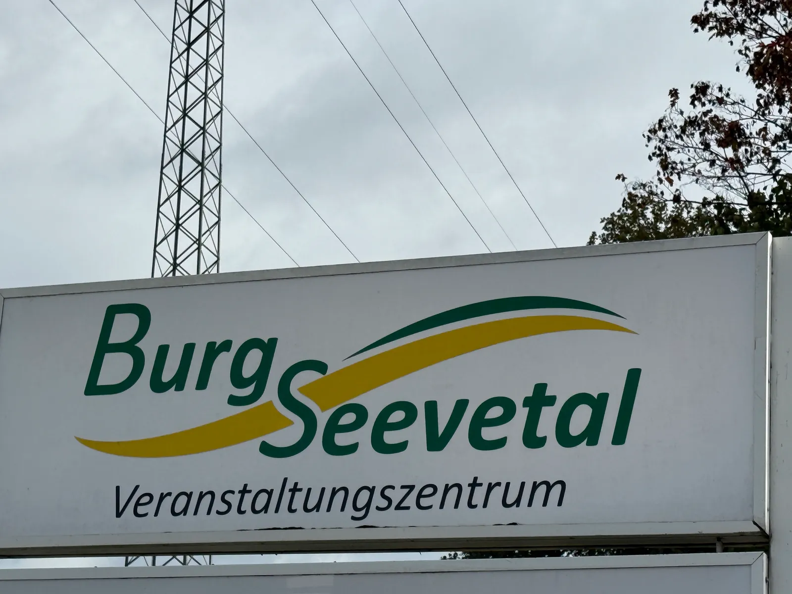 Schild mit grün-gelbem Schriftzug „Burg Seevetal Veranstaltungszentrum“, dahinter Gittermast unter grauem Himmel