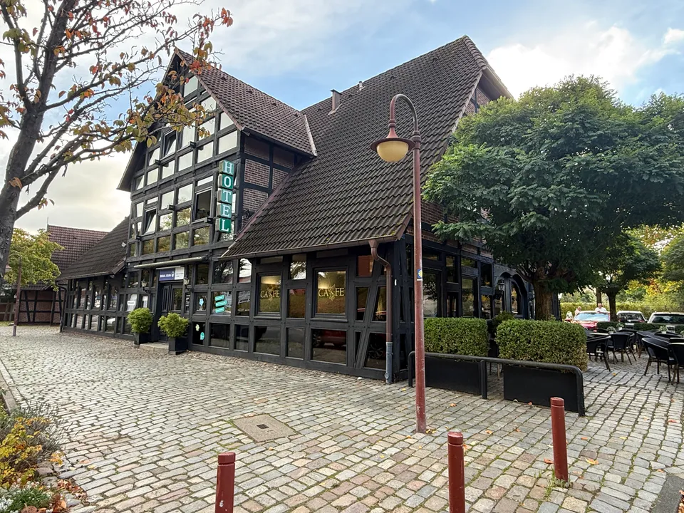 Fachwerkgebäude mit senkrechtem grünem Hotel-Schild und Café-Fenstern, davor Kopfsteinpflaster, Straßenlaterne und rotbraune Poller