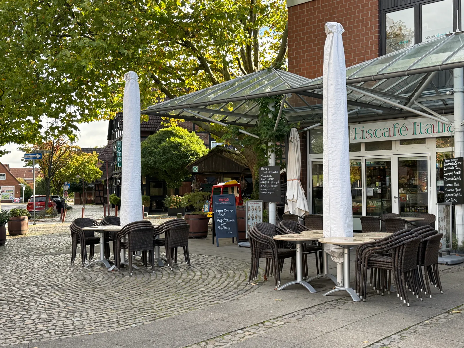 Terrasse des Eiscafé Italia mit gestapelten Rattanstühlen, verhüllten Sonnenschirmen und Glasvordach vor einem Backsteingebäude