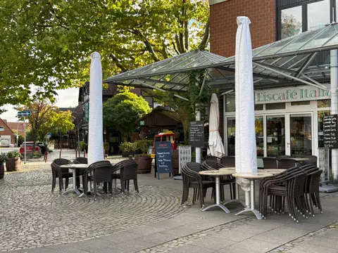 Terrasse des Eiscafé Italia mit gestapelten Rattanstühlen, verhüllten Sonnenschirmen und Glasvordach vor einem Backsteingebäude