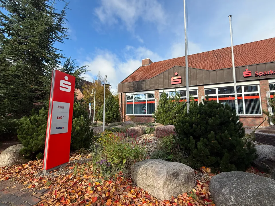 Rote Sparkassen-Stele mit Deka-, LBS- und VGH-Logos vor Backsteingebäude, Steingarten mit Herbstlaub