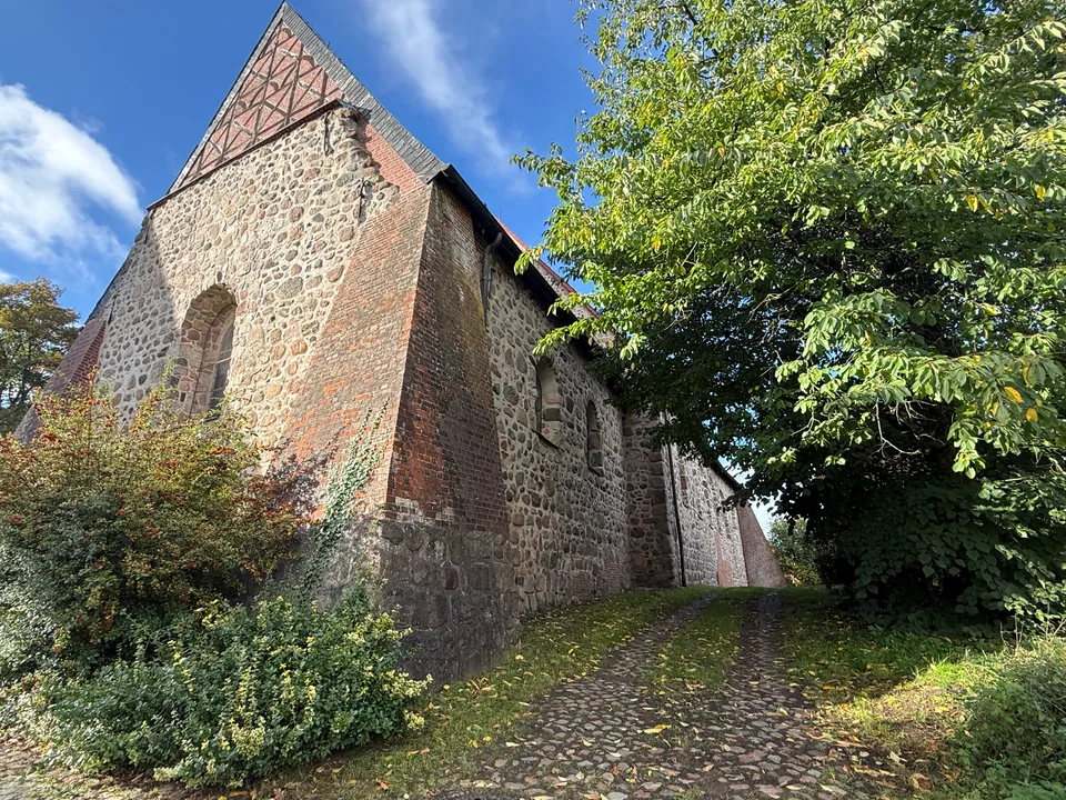 Giebel und Seitenwand einer Feldsteinkirche mit Backsteinstreben, daneben ein Pflasterweg unter einem Laubbaum
