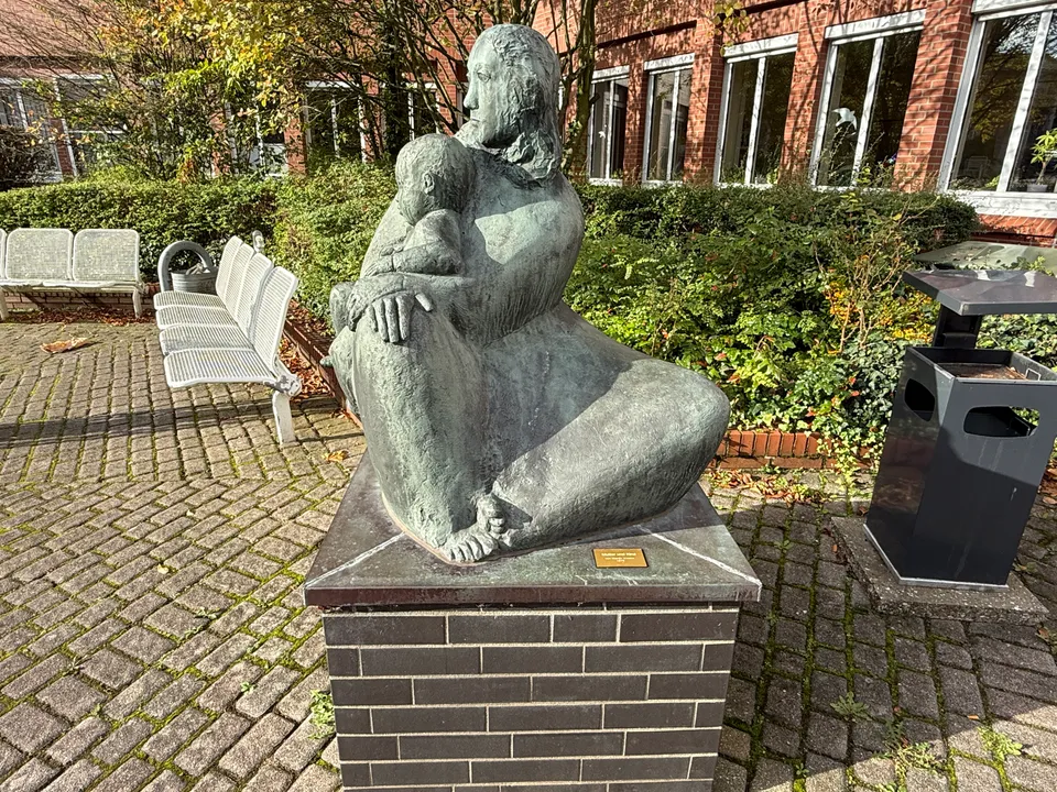 Bronzeskulptur einer sitzenden Frau mit Kind auf einem Klinkersockel, dahinter Bänke und eine Klinkerfassade