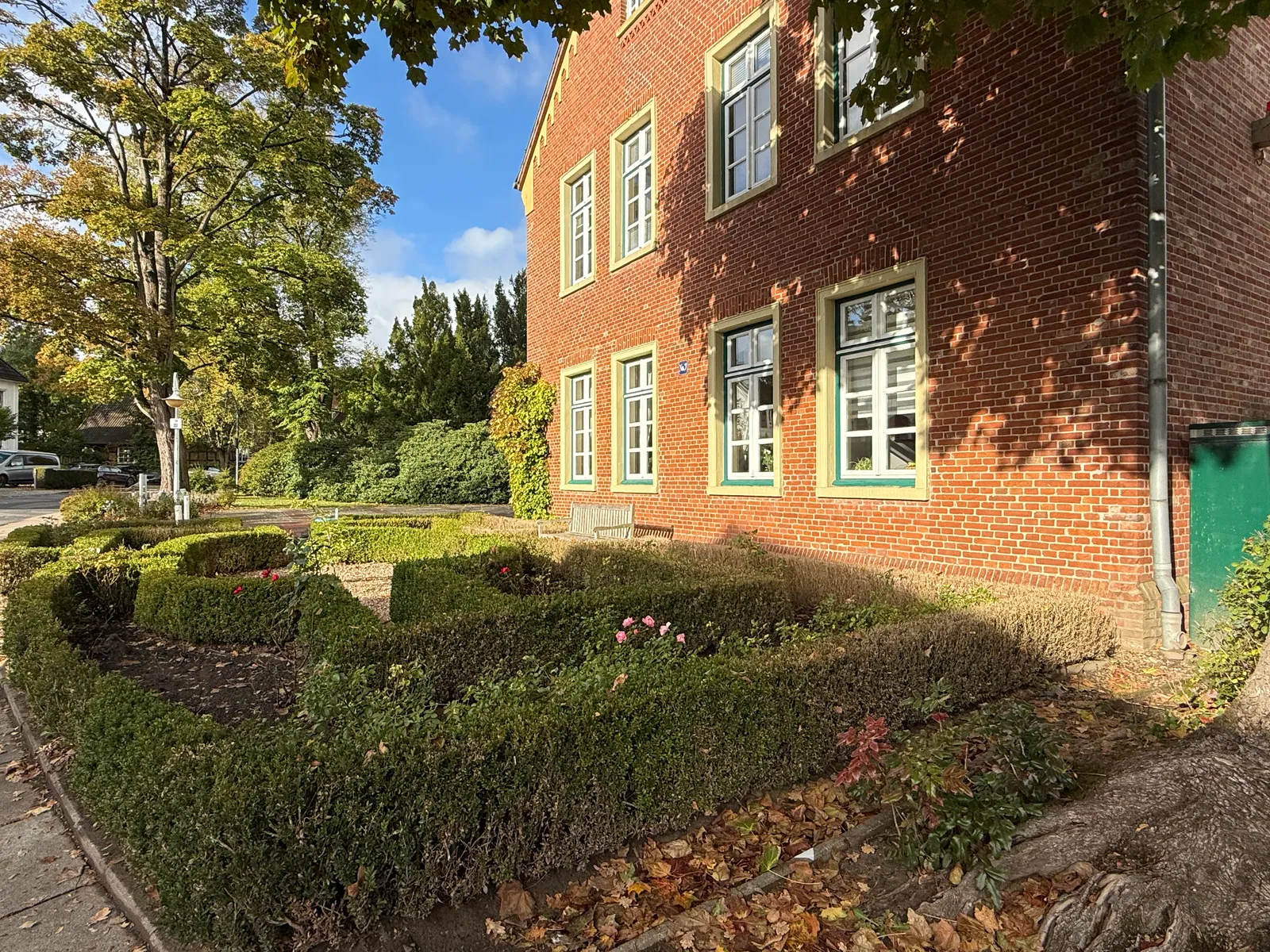 Seitenfassade eines roten Backsteingebäudes mit Ziergarten aus Buchsbaumhecken, Rosen und Holzbank im Herbstlicht