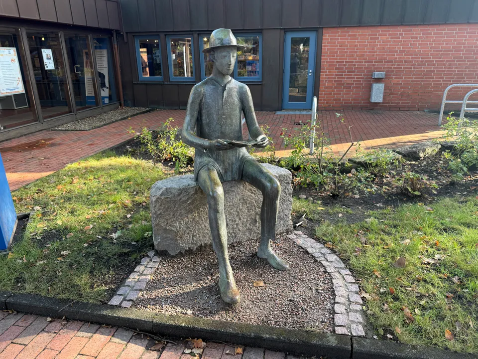 Bronzeskulptur einer lesenden Figur mit Hut auf einem Granitblock vor einer Klinkerfassade mit blauen Fenstern