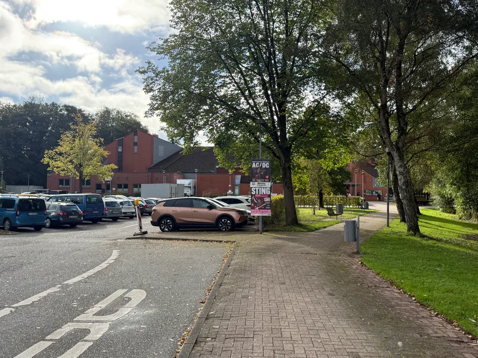 Parkplatz und gepflasterter Fußweg mit Bäumen vor rotem Klinkergebäude, am Mast hängen Konzertplakate