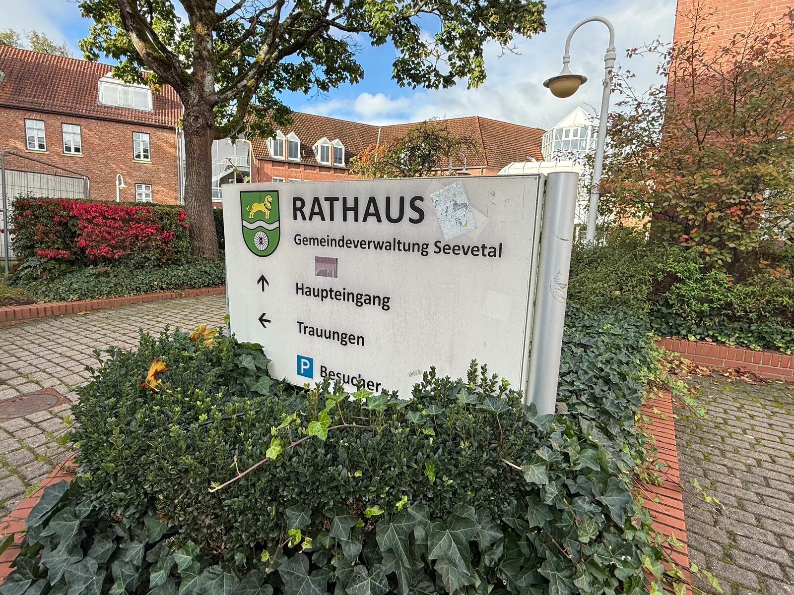 Wegweiser „Rathaus Gemeindeverwaltung Seevetal“ mit Pfeilen zu Haupteingang, Trauungen und Besucherparkplatz in einem Efeubeet
