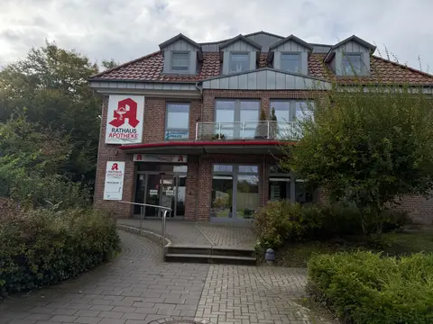 Backsteingebäude der Rathaus Apotheke Hittfeld mit rotem Apotheken-A, Vordach, Glaseingang und gepflastertem Zugangsweg zwischen Hecken