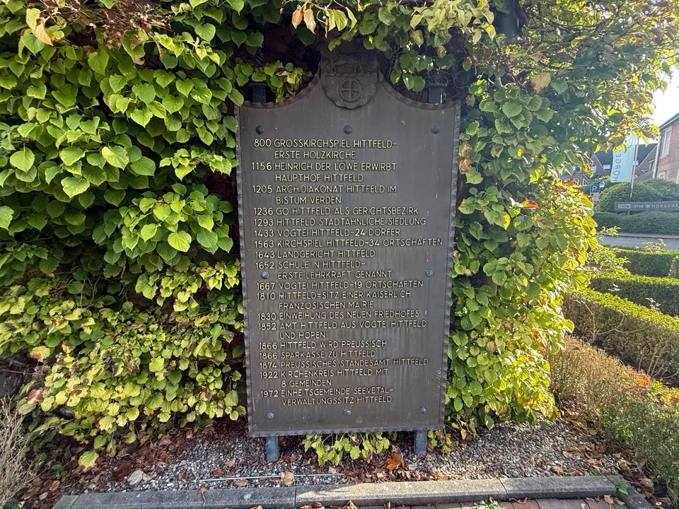 Metallene Chroniktafel mit Jahreszahlen zur Geschichte Hittfelds von 800 bis 1972 vor einer berankten Wand
