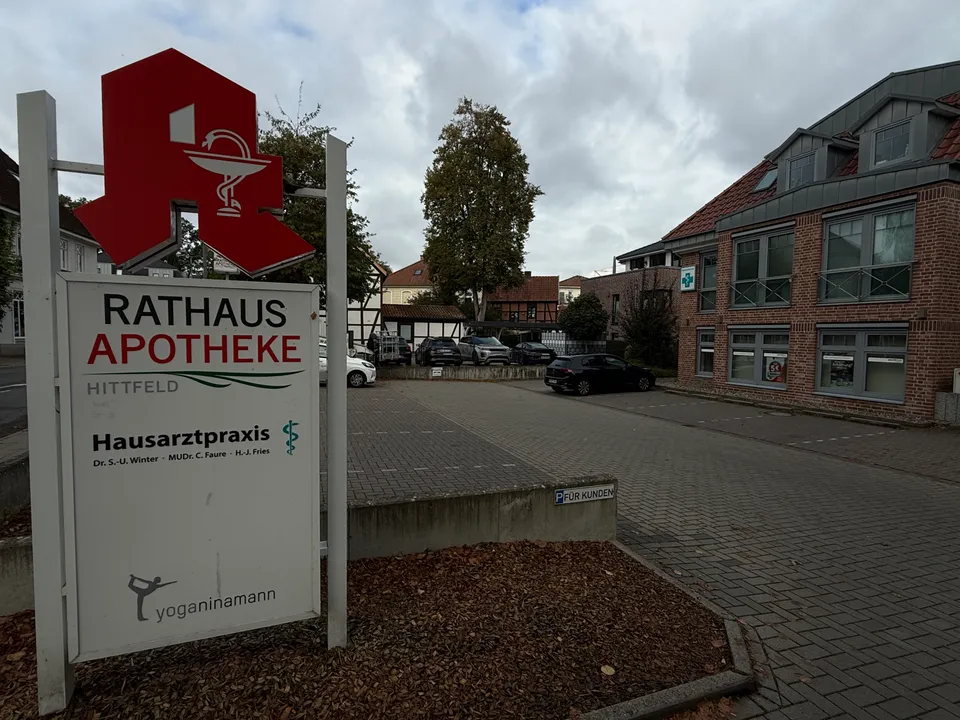 Freistehendes Schild der Rathaus Apotheke Hittfeld vor einem gepflasterten Kundenparkplatz, Backsteingebäude und Fachwerkhäusern im Hintergrund