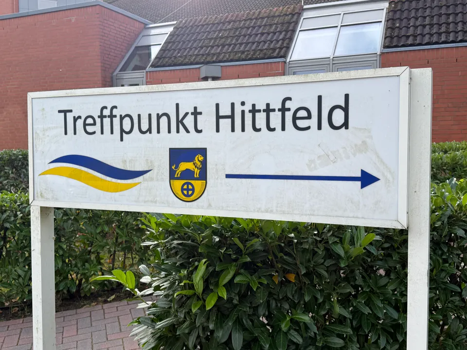 Weißer Wegweiser „Treffpunkt Hittfeld“ mit Löwenwappen und Pfeil nach rechts vor Hecke und Klinkergebäude