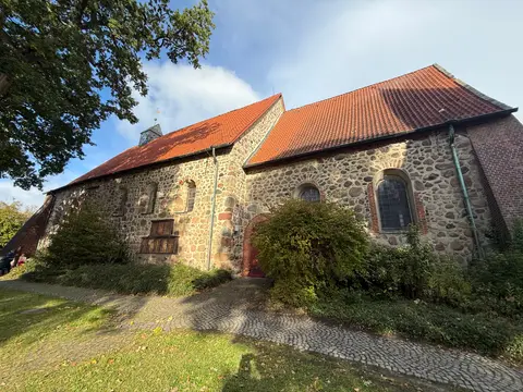 Längsseite einer Feldsteinkirche mit rotem Ziegeldach, Rundbogenfenstern, Pflasterweg und Rasen im Sonnenlicht