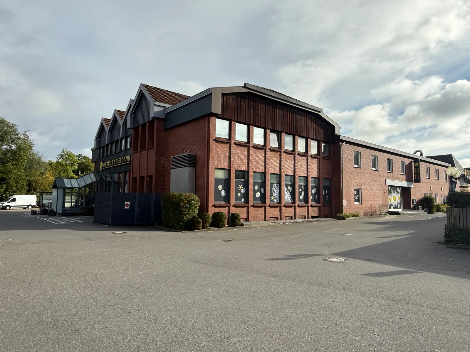 Langgestreckter Klinkergebäudekomplex mit Schriftzug „Merkur Spielbank“, davor ein großer leerer Asphaltparkplatz unter wolkigem Himmel