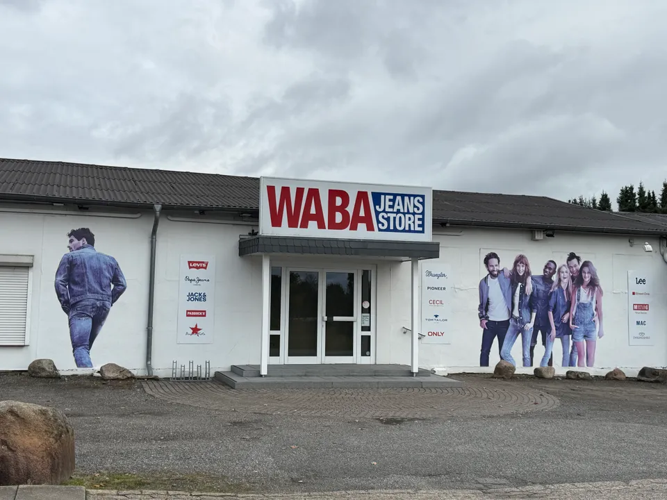Weißes Flachdachgebäude mit Schild „WABA Jeans Store“, Jeanswerbung an der Fassade, Parkplatz mit Findlingen davor