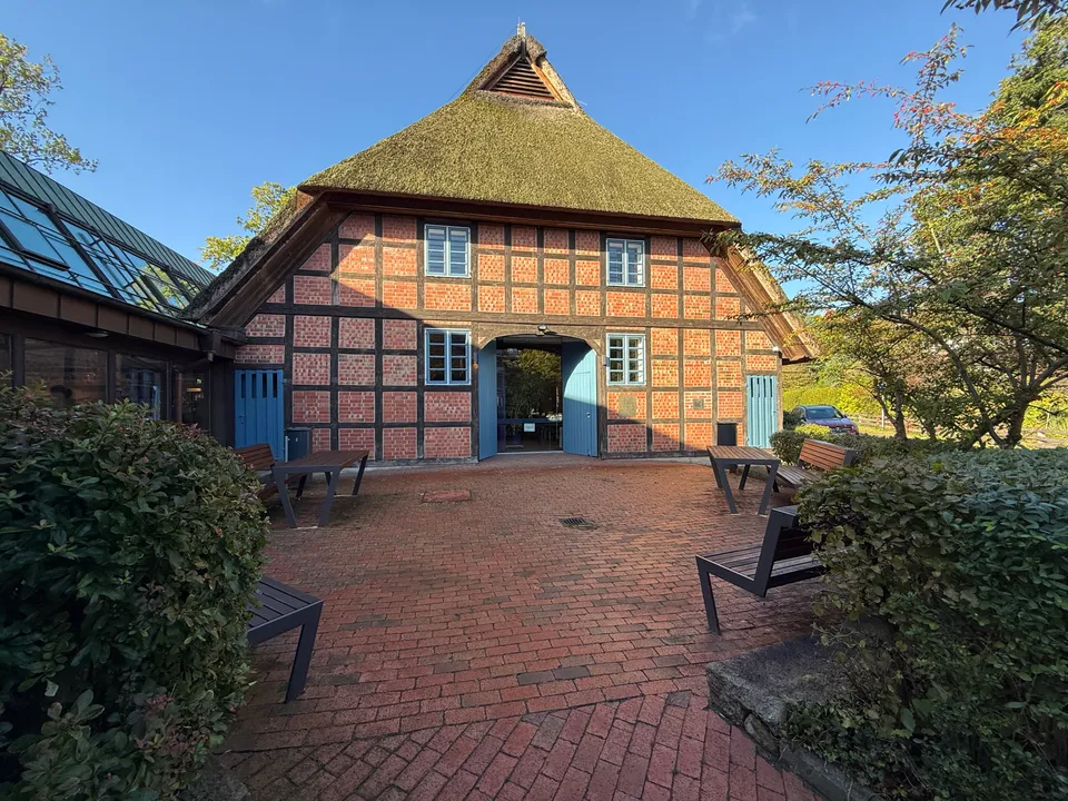 Reetgedecktes Fachwerkhaus mit offenem blauem Tor, davor gepflasterter Platz mit Bänken und Tischen