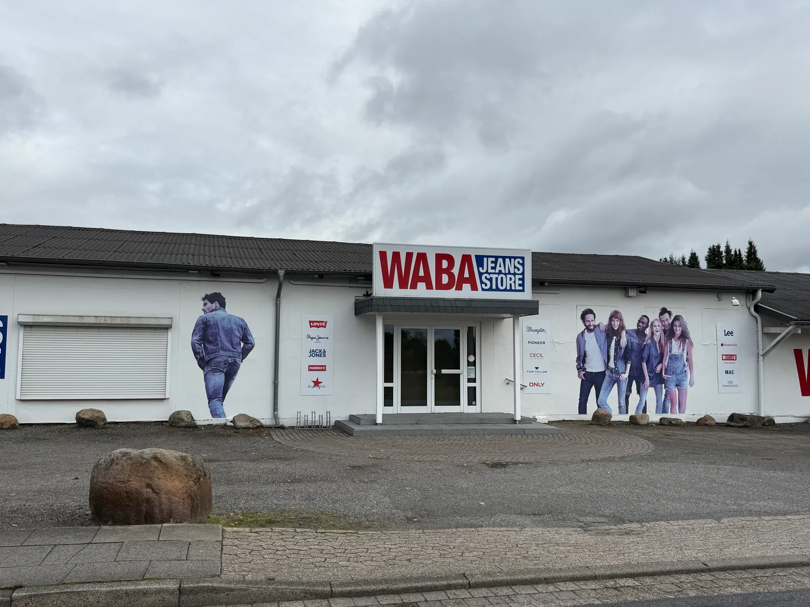 Jeansgeschäft mit rot-blauem Schild „WABA Jeans Store“ an weißer Fassade, Findling und Gehweg im Vordergrund