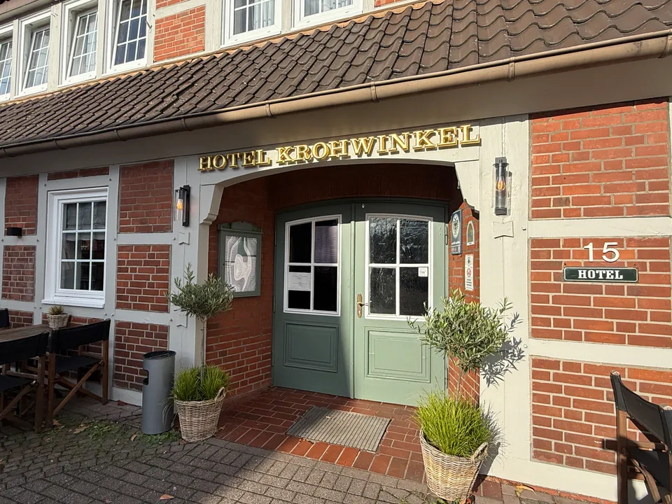 Eingang des Hotels Krohwinkel mit grüner Holztür, goldenem Schriftzug, Fachwerkfassade und Olivenbäumchen in Körben