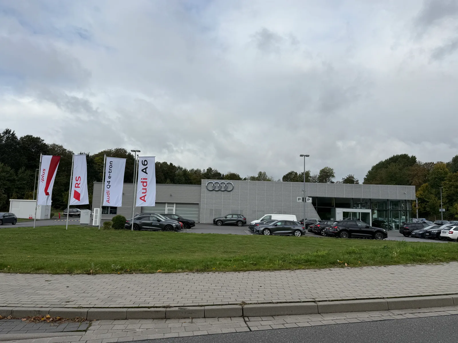 Audi-Autohaus mit grauer Metallfassade, Werbefahnen und geparkten Fahrzeugen hinter einer Rasenfläche unter bewölktem Himmel