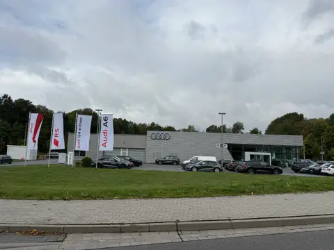 Audi-Autohaus mit grauer Metallfassade, Werbefahnen und geparkten Fahrzeugen hinter einer Rasenfläche unter bewölktem Himmel