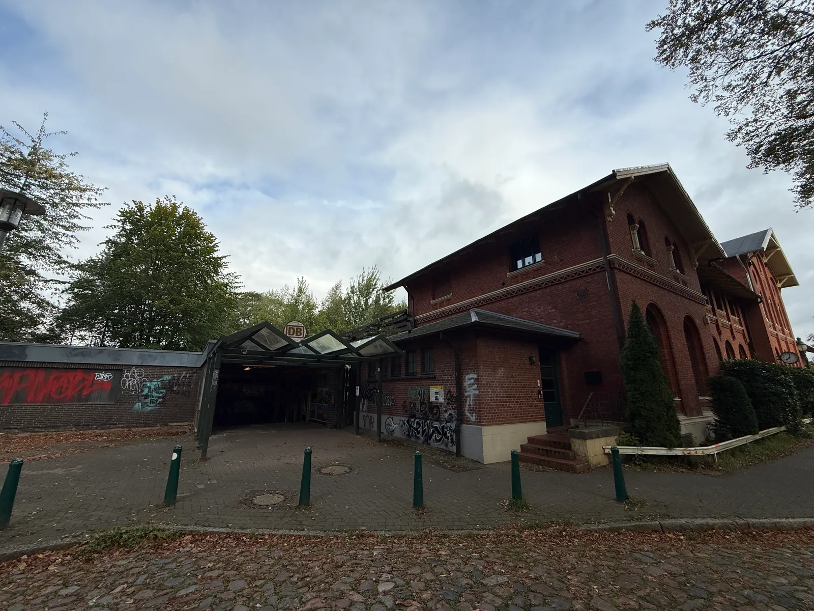 Rotes Backstein-Bahnhofsgebäude mit überdachtem Zugang, DB-Logo, grünen Pollern und Graffiti an der Mauer