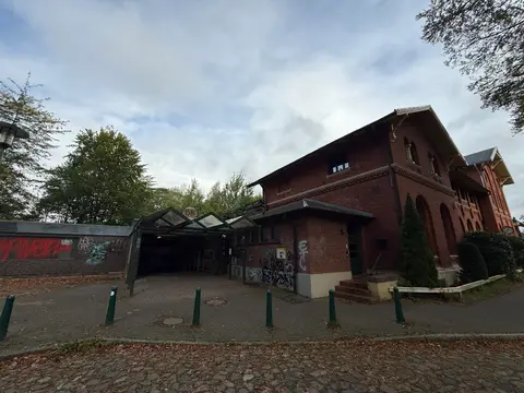Rotes Backstein-Bahnhofsgebäude mit überdachtem Zugang, DB-Logo, grünen Pollern und Graffiti an der Mauer