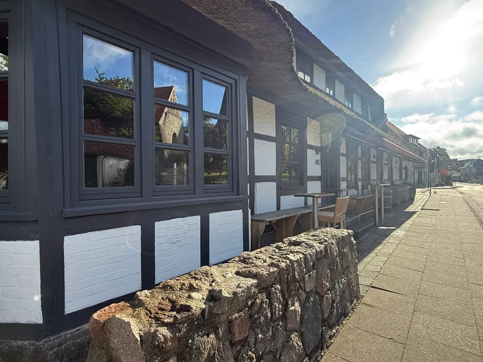 Fachwerkhaus mit Reetdach und grauen Sprossenfenstern hinter Feldsteinmauer, daneben Gehweg im Gegenlicht