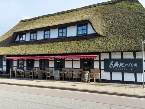 Reetgedecktes Fachwerkhaus mit roten Markisen, Fass-Stehtischen, Barhockern und Schild „BARrica hittfeld“ an der Straße