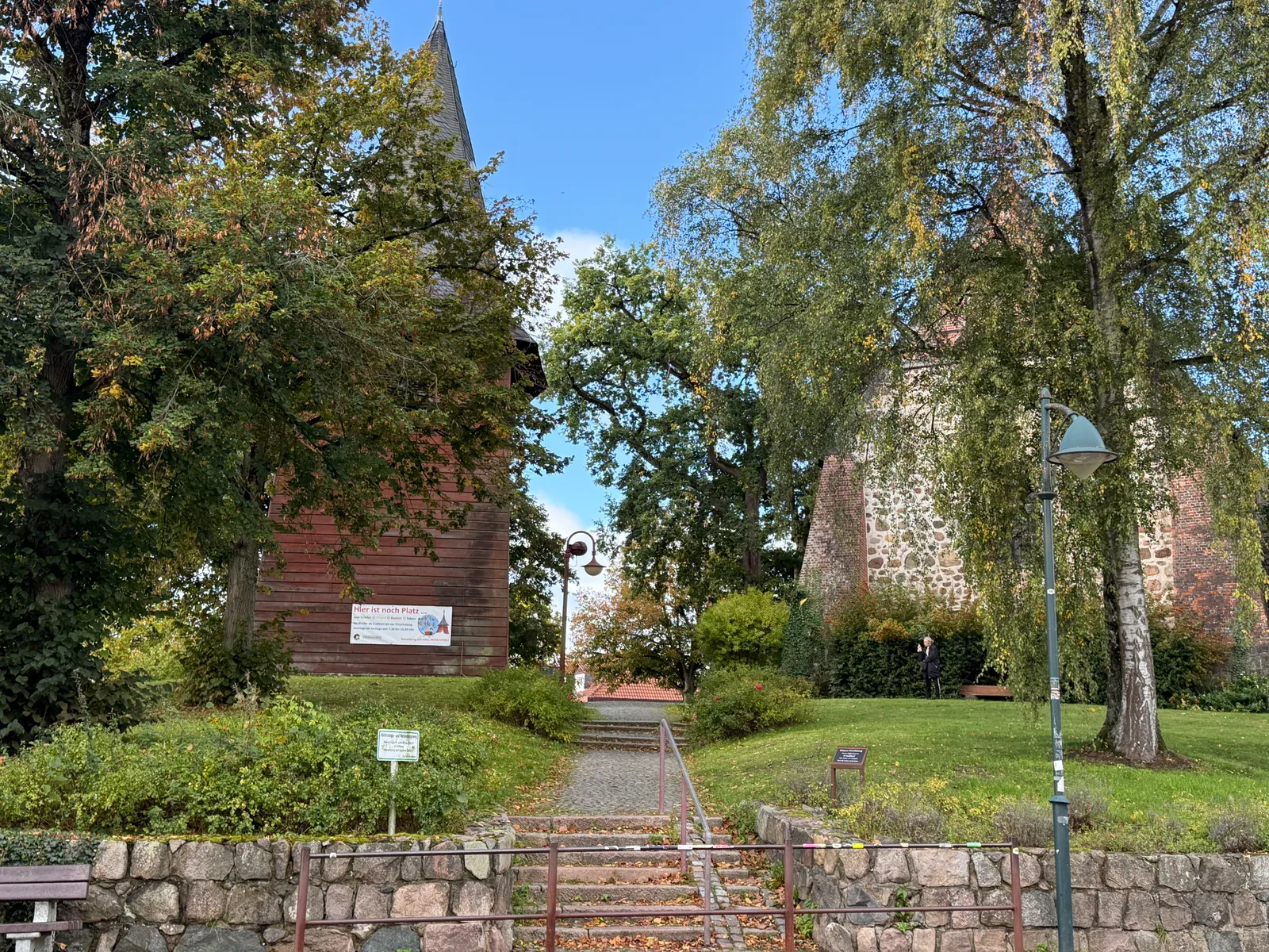 Treppe mit Geländer führt zu einem roten Holzglockenturm und einer Feldsteinkirche zwischen Bäumen