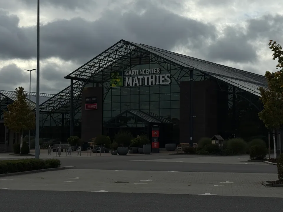 Dunkel belichtete Frontansicht des Gartencenters Matthies mit Glasfassade, Eingangsbereich und leerem Parkplatz unter Wolken