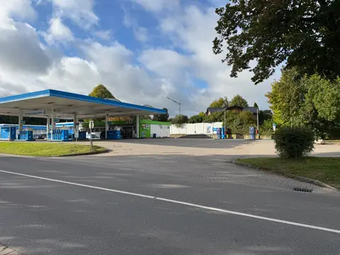 Tankstelle mit blauem Tankdach, Zapfsäulen und grünem REWE-to-GO-Shop hinter einer gepflasterten Zufahrt an einer Straße
