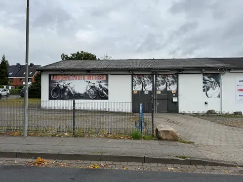 Weiße Gewerbehalle mit großem Banner „Fahrrad-Lager-Hittfeld“, Holztor, Zaun und Fahrradständern bei bedecktem Himmel