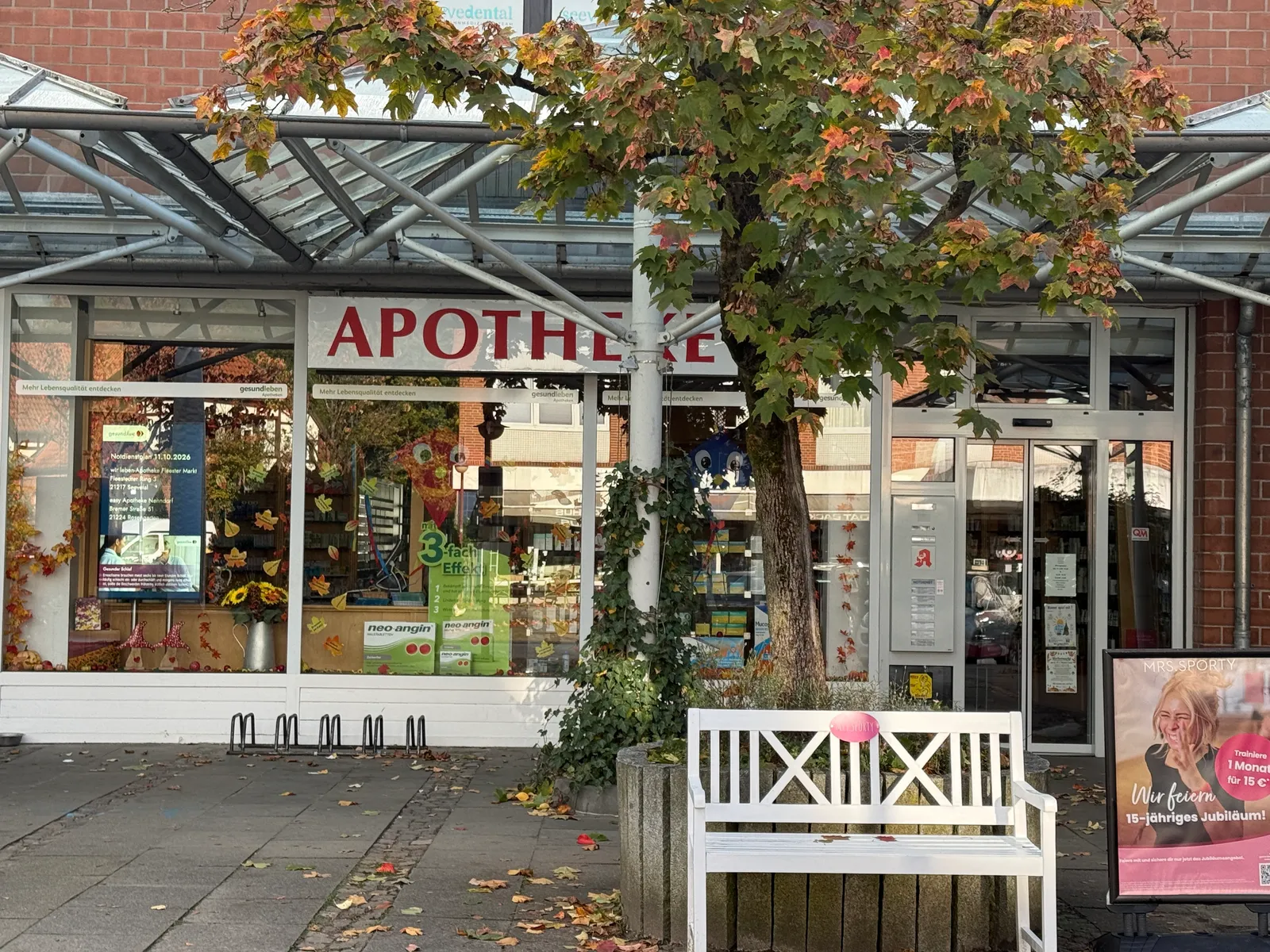 Apotheke mit Glasvordach und herbstlich dekorierten Schaufenstern, davor Baum mit buntem Laub und weiße Holzbank