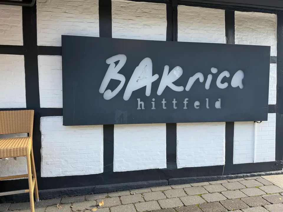 Dunkles Schild mit heller Aufschrift „BARrica hittfeld“ an weiß-schwarzer Fachwerkwand