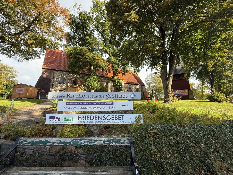 Hinweisschilder der Kirchengemeinde Hittfeld vor einer Feldsteinkirche zwischen Bäumen, vorne eine alte Parkbank