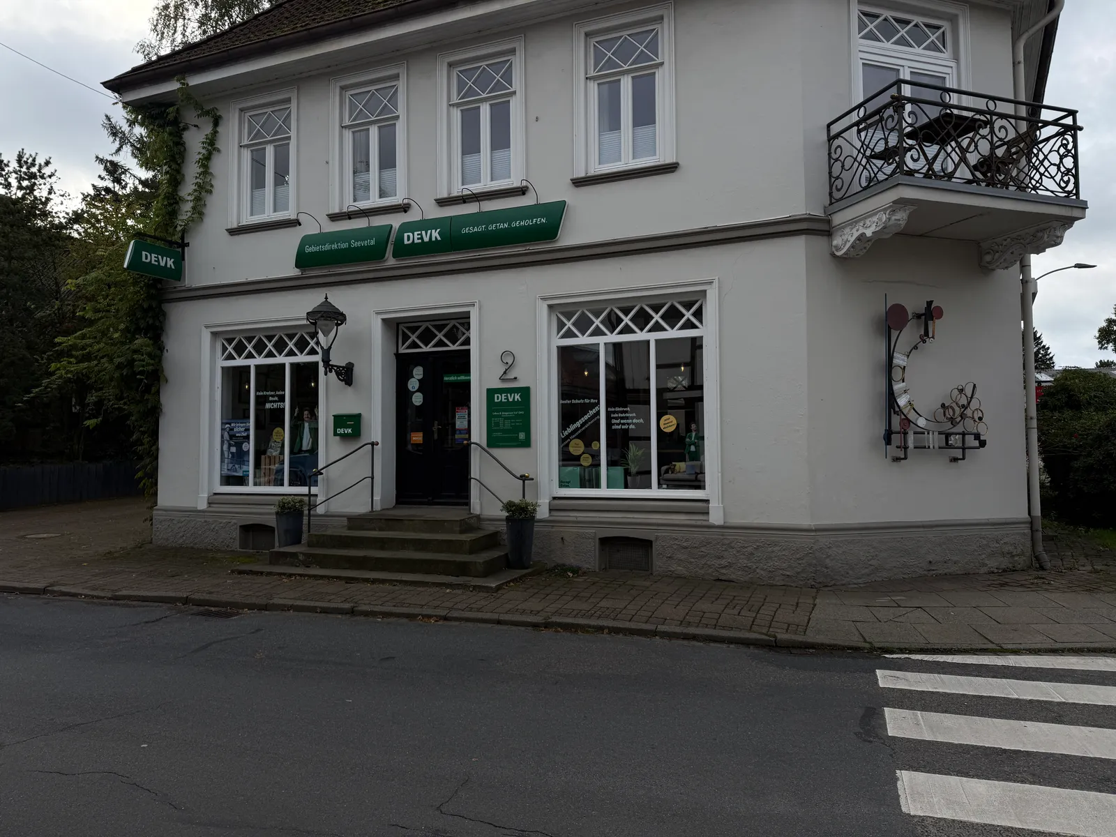 Helles Eckhaus mit DEVK-Geschäftsstelle, Schaufenstern, Steintreppe zum Eingang, Eckbalkon und Zebrastreifen im Vordergrund