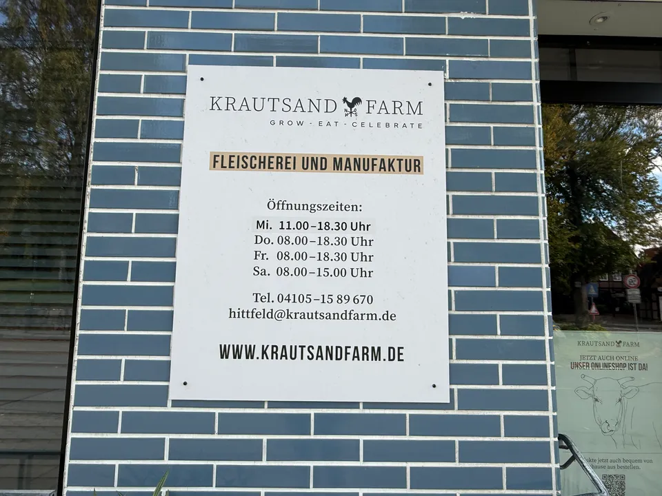 Weißes Schild der Krautsand Farm mit Öffnungszeiten und Kontaktdaten an blauer Klinkerwand neben einem Schaufenster