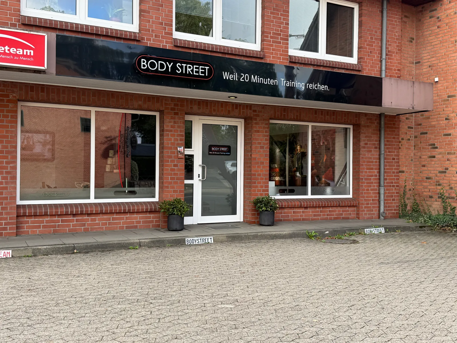 Klinkerfassade mit schwarzem Schild „Body Street“, Glastür, zwei Schaufenstern, Pflanzkübeln und gepflastertem Parkplatz davor