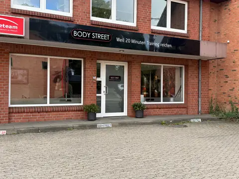 Klinkerfassade mit schwarzem Schild „Body Street“, Glastür, zwei Schaufenstern, Pflanzkübeln und gepflastertem Parkplatz davor