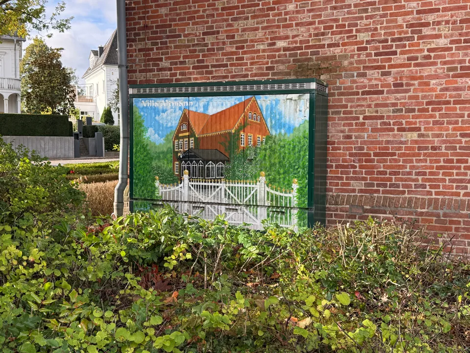 Bemalter Verteilerkasten mit Darstellung einer historischen Villa mit weißem Zaun vor einer Ziegelwand