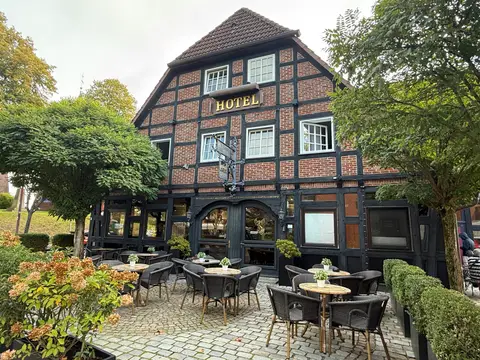 Fachwerkhaus aus rotem Backstein mit goldenem Hotel-Schriftzug am Giebel, davor gepflasterte Terrasse mit Rattanstühlen und Tischen