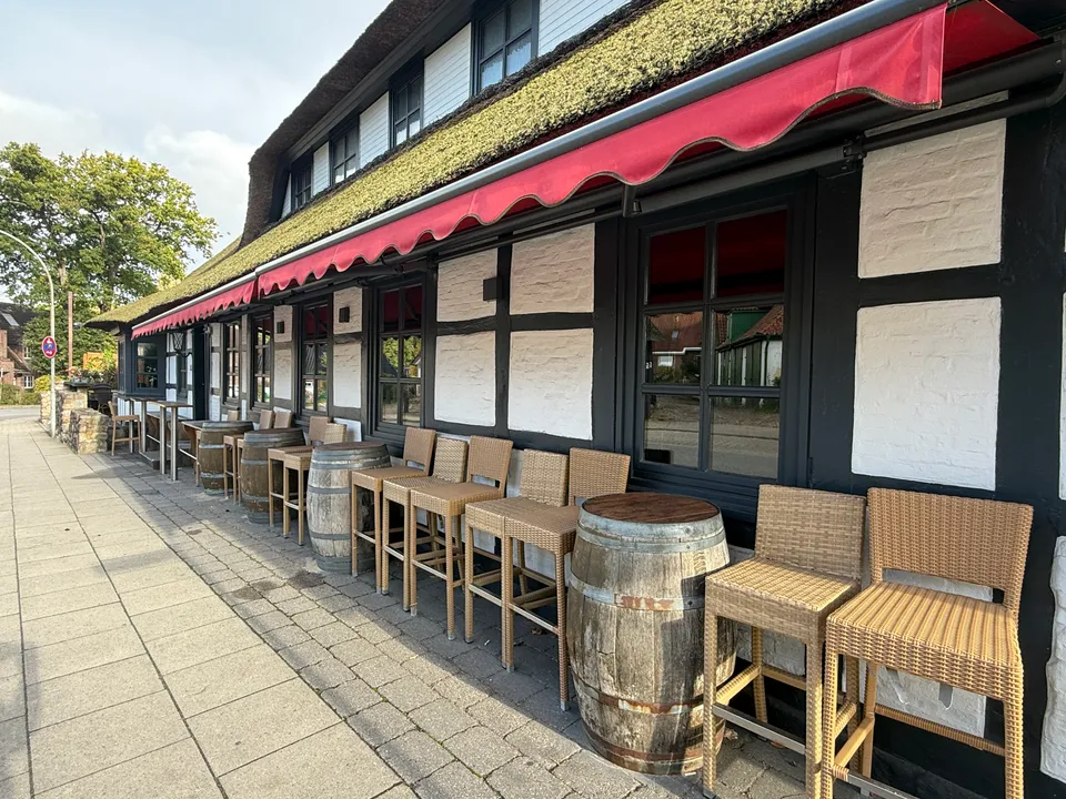 Reihe aus Rattan-Barhockern und Holzfässern unter roter Markise entlang einer Fachwerkfassade mit Reetdach