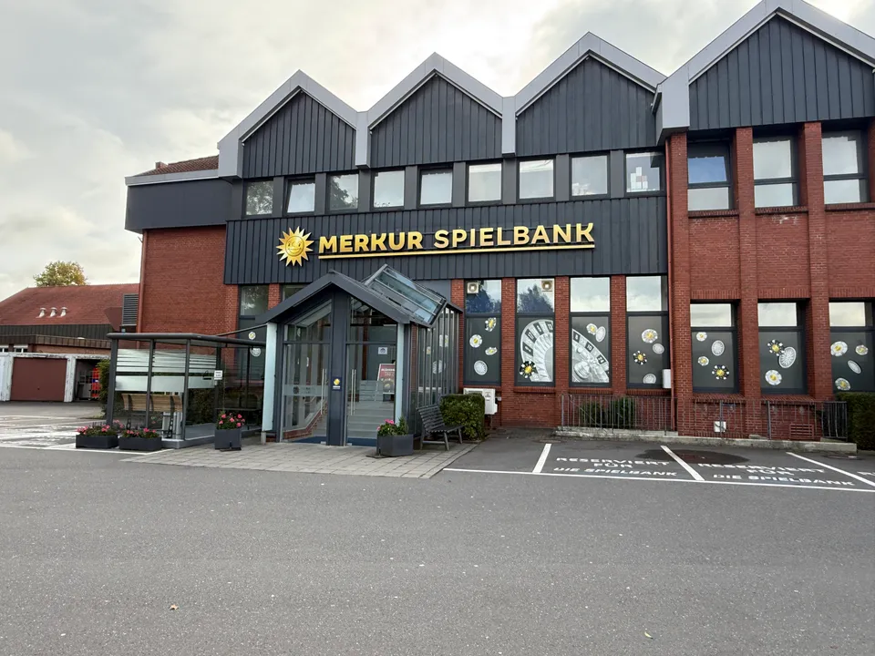 Klinkergebäude mit dunklen Giebeln, goldenem Schriftzug „Merkur Spielbank“, verglastem Eingang und reservierten Parkplätzen davor