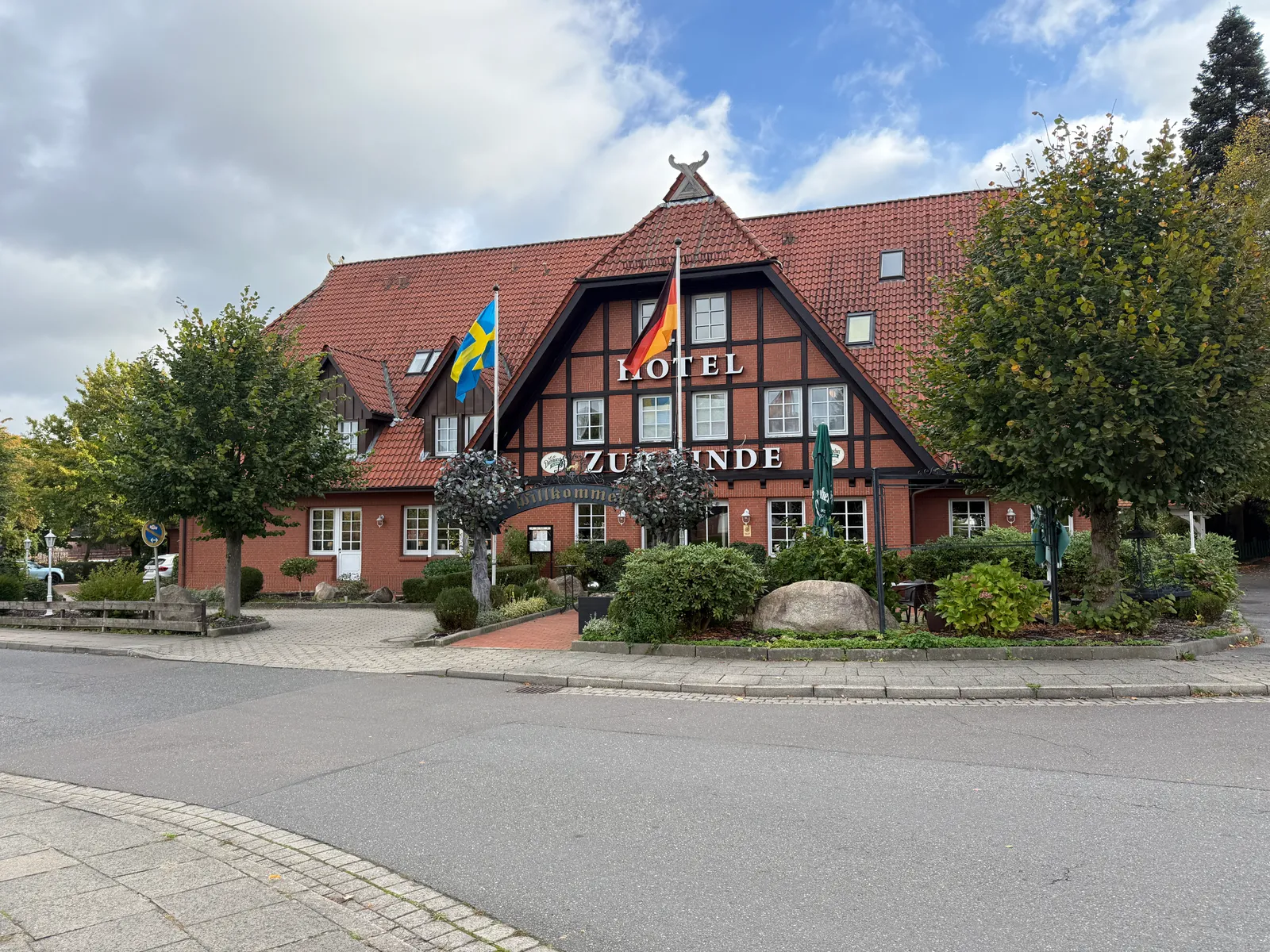 Rotes Fachwerkhotel Zur Linde mit Flaggen und Bäumen, von der gegenüberliegenden Straßenseite aus gesehen
