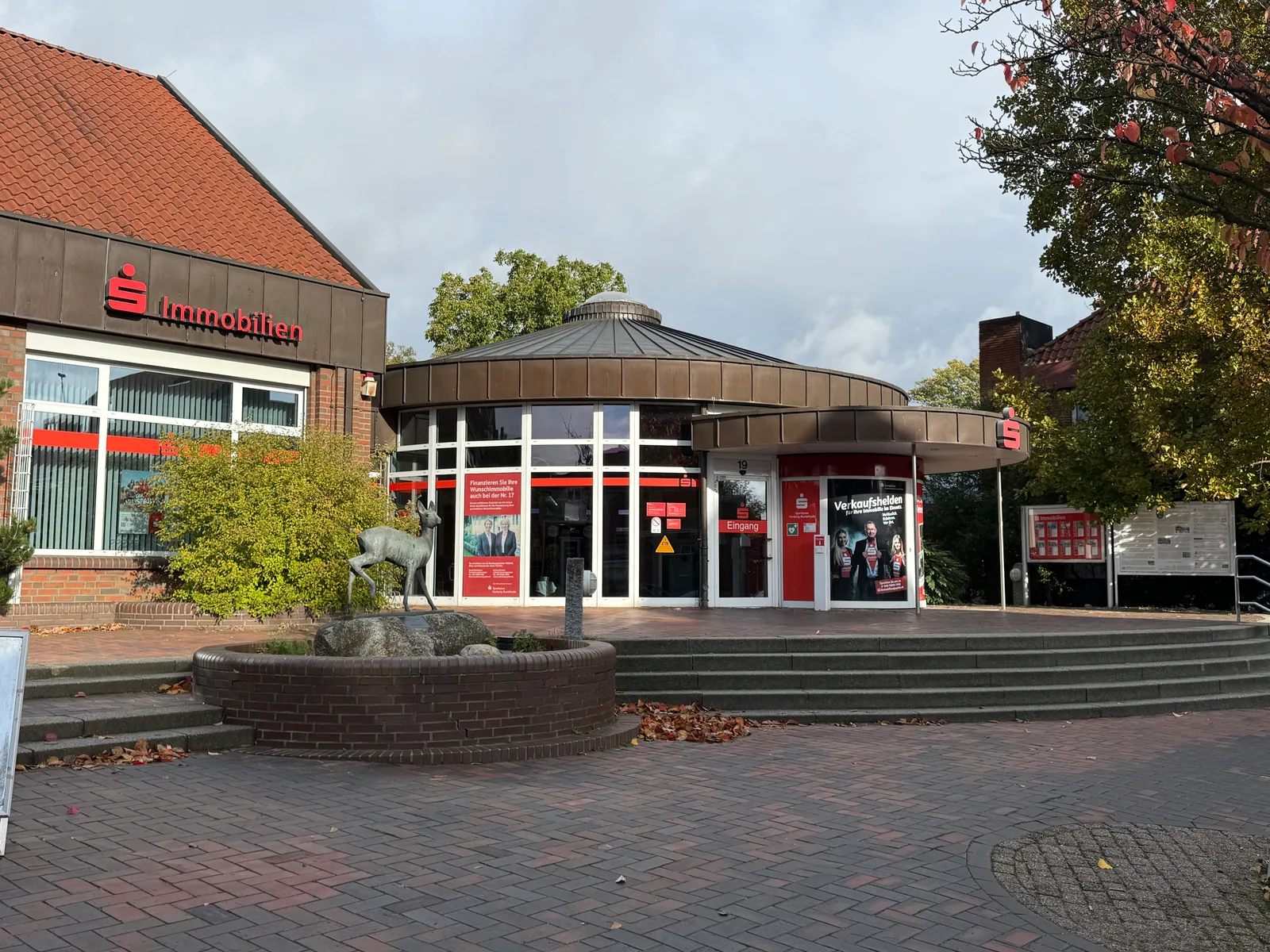 Runder Sparkassen-Pavillon mit Glasfront, davor gepflasterter Platz, Stufen und Backsteinbrunnen mit Rehskulptur