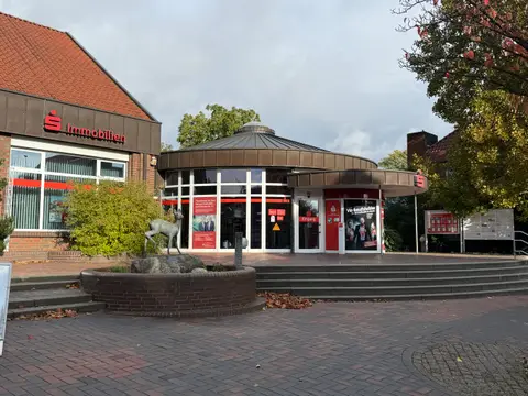 Runder Sparkassen-Pavillon mit Glasfront, davor gepflasterter Platz, Stufen und Backsteinbrunnen mit Rehskulptur