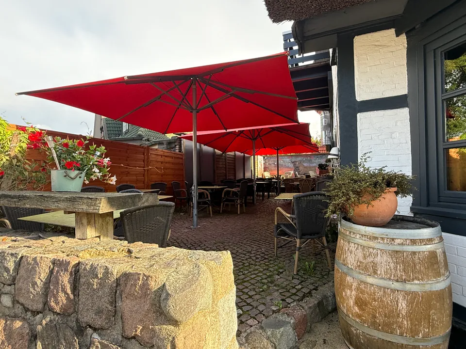 Gepflasterter Biergarten mit roten Sonnenschirmen, Geflechtstühlen und Holzfass hinter einer Feldsteinmauer
