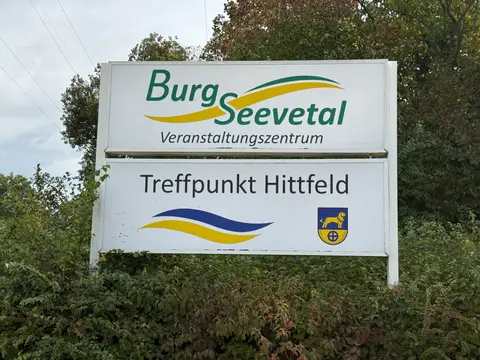 Weißes Schild „Burg Seevetal Veranstaltungszentrum“ und „Treffpunkt Hittfeld“ mit Wappen vor grünem Gebüsch