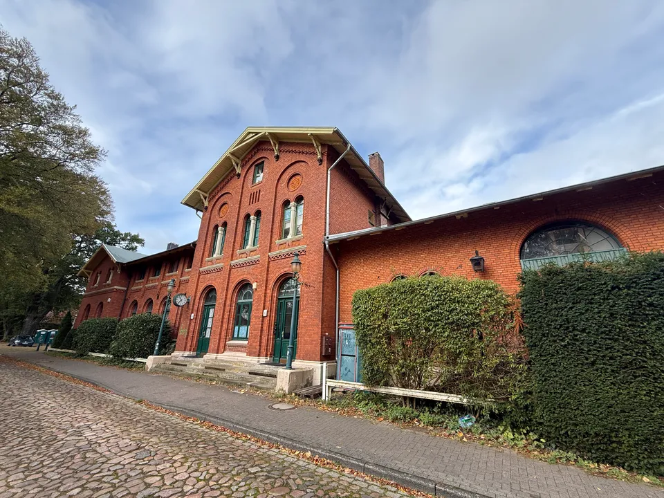 Historisches rotes Backstein-Bahnhofsgebäude mit Ziergiebel, Rundbogenfenstern, Uhr und Hecken an gepflasterter Straße